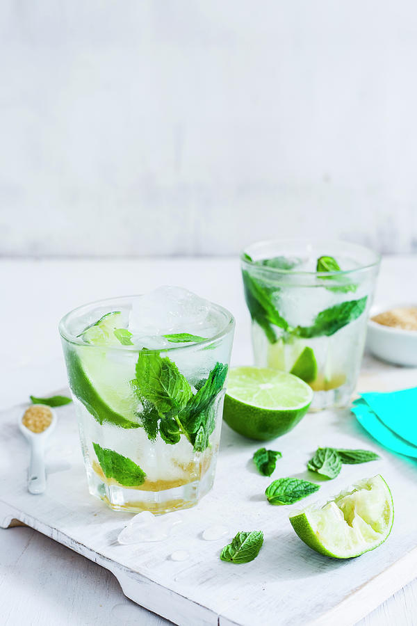 Two Glasses With Classic Mojito, Lime Wedges And Mint Leaves On A White Wood Cutting Board Photograph by Maricruz Avalos Flores