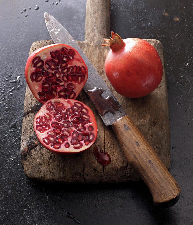 Two Pomegranates, Whole And Halved, With A Knife On An Old Wooden Board Photograph by Ludger Rose