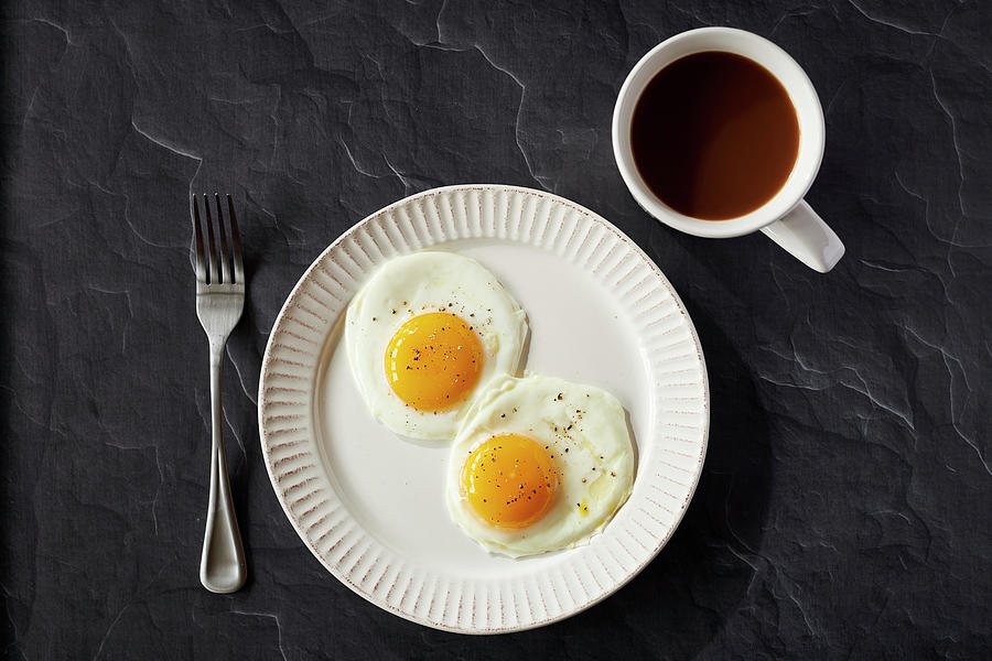 Two Sunnyside Eggs With Pepper In A White Plate With Coffee In A White Coffee Cup On A Black Surface Photograph by Jillian Graniero