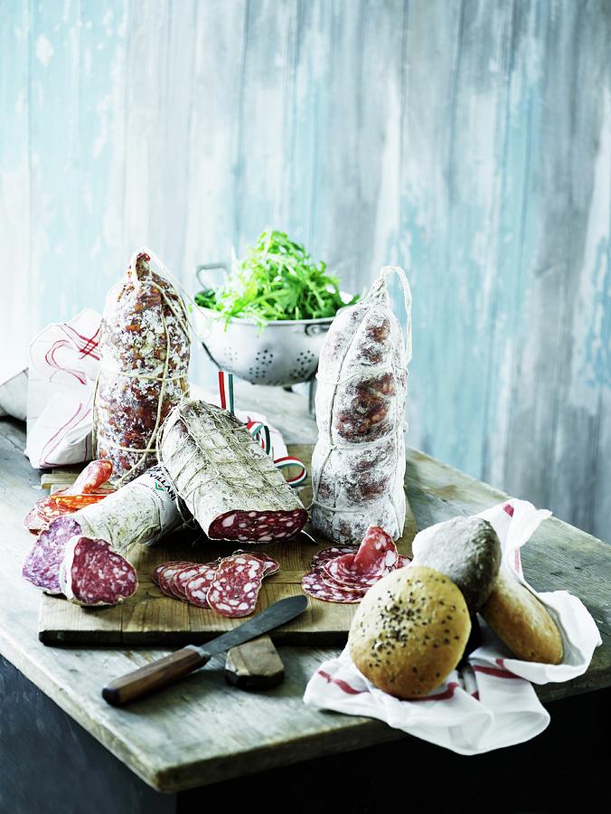 Various Types Of Salami On A Wooden Chopping Board With Roles And Freshly Washed Lettuce Photograph by Mikkel Adsbl