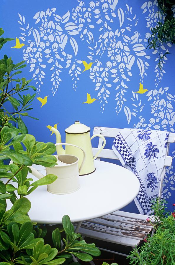 Wall Deco With Humming Birds And Wisteria On Blue-painted Garden Wall Behind White Table, Chair And Coffee Pot Photograph by Linda Burgess