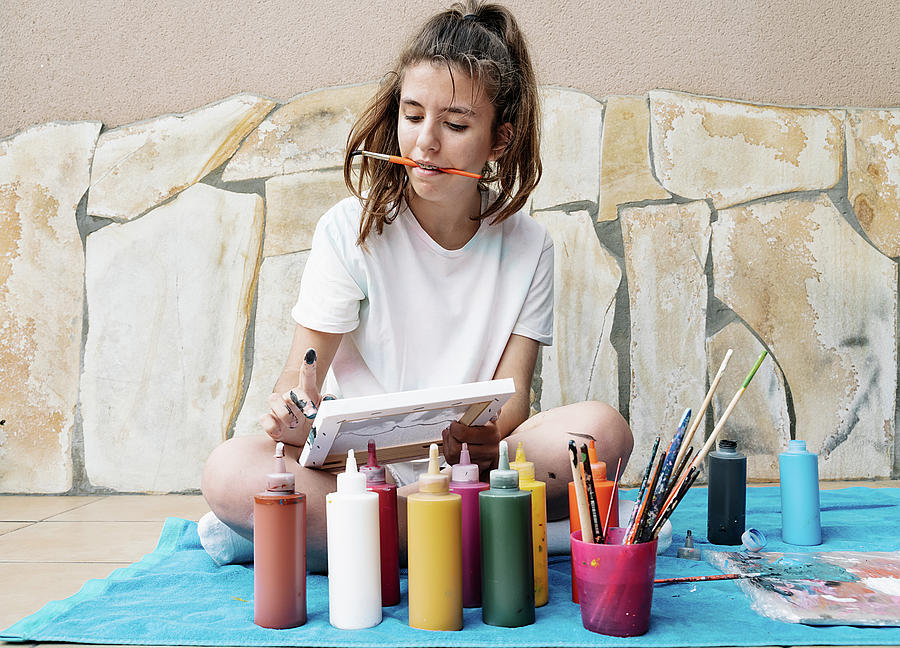 Abstract Photograph - White Girl Biting A Painting Brush While Painting With Her Finger Her Picture Siting On A Terrace In Front Of Bottle Paintings. Horizontal Photo by Cavan Images
