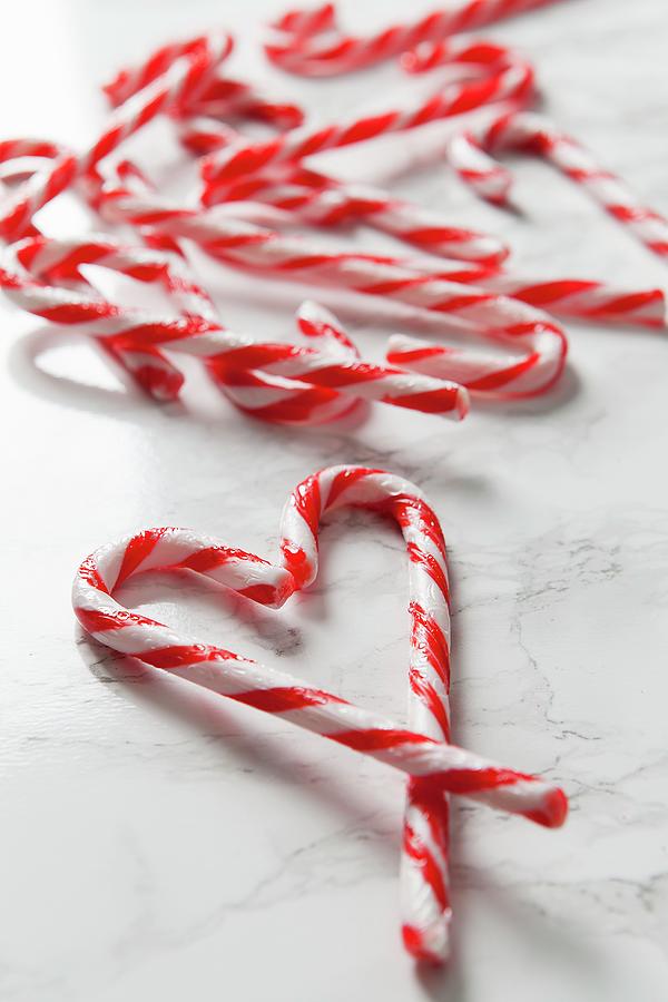 White Marble Surface Covered In Candy Canes And Two Canes In The Foreground Forming A Heart Shape Photograph by Stacy Grant