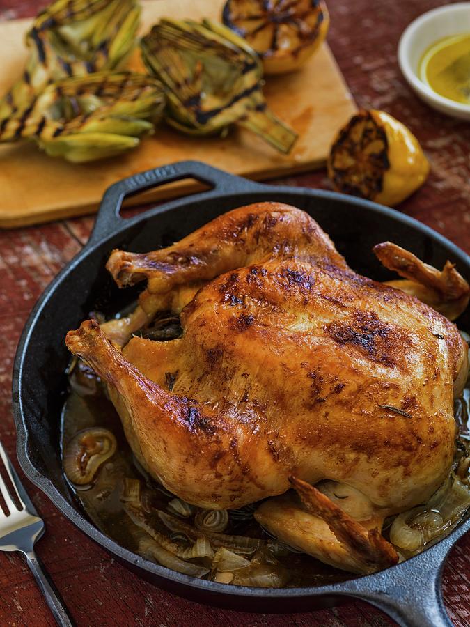 Whole Roasted Chicken In A Cast Iron Pan On A Rustic Surface With Grilled Baby Artichokes And Lemon In The Background Photograph by Don Crossland