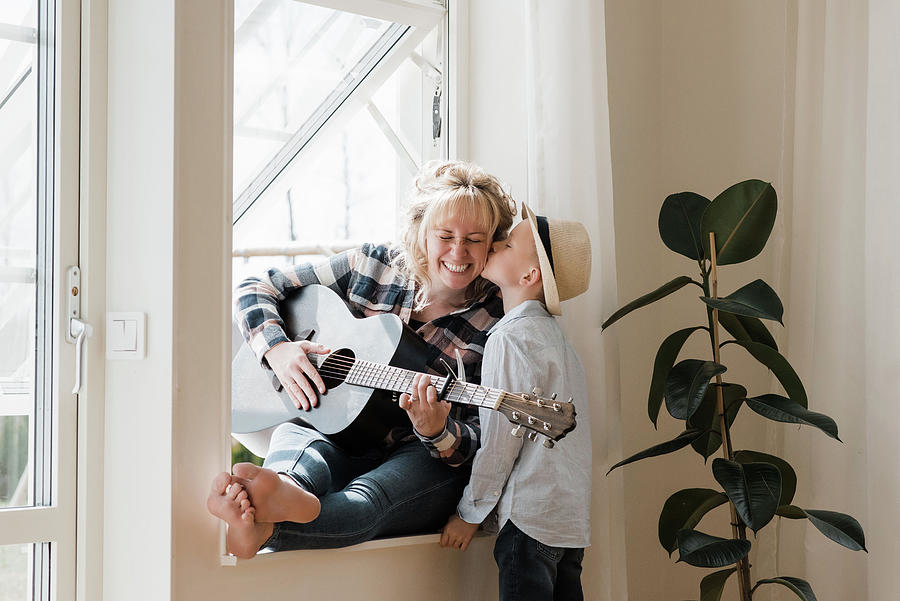 Parenthood Movie Photograph - Woman Sat Playing Guitar At Home Whilst Her Son Gives Her A Kiss by Cavan Images