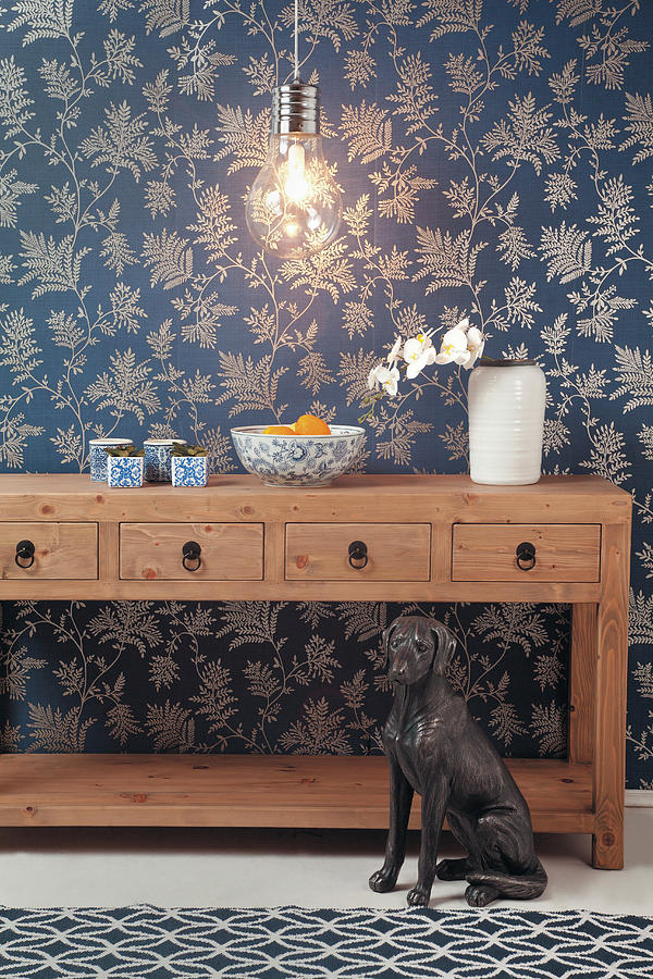 Wooden Console Table With Drawers, Light Bulb Lamp And Dog Sculpture Against Blue Floral Wallpaper Photograph by Great Stock!