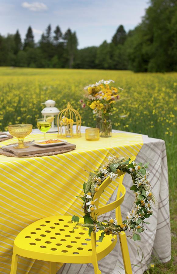 Wreath Of Flowers On Yellow Metal Chair Next To Table Set In Yellow Colour Scheme With Field Of Flowering Rapeseed In Background Photograph by Annette Nordstrom