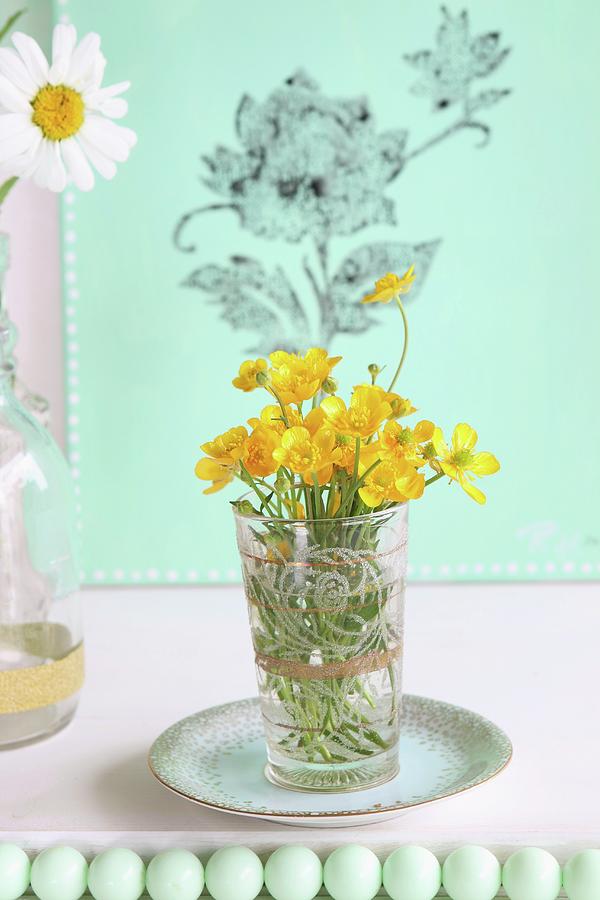 Yellow Wildflowers In Glass On Saucer In Front Of Green Canvas Printed With Floral Pattern; Row Of Green Beads In Foreground Photograph by Regina Hippel
