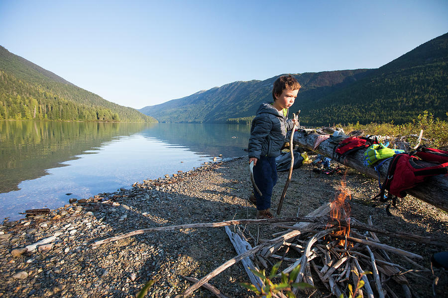 Nature Photograph - Young Boy Carries Sticks To Add To Lakeside Fire During A Camping Trip by Cavan Images