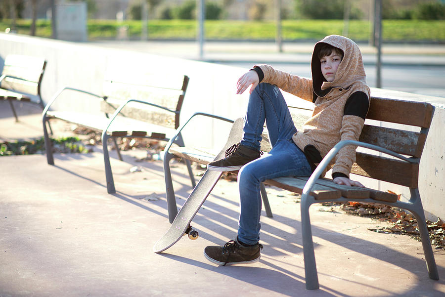 Sunset Photograph - Young Hooded Boy Sitting On Bench Next His Skateboard, Looking Camera by Cavan Images