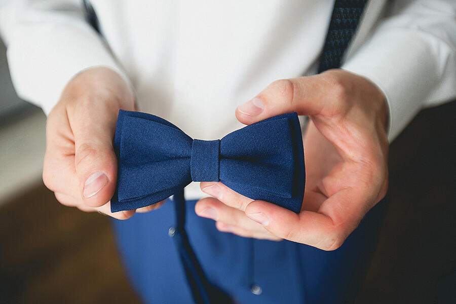 A man, a groom or a businessman in a stylish suit holding a blue bow tie, a fashion accessory. Preparing the groom for the wedding day. Blurred background, close-up. #1 Photograph by Aleksandr Zubkov