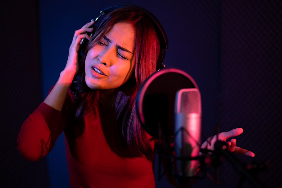 Beautiful young woman singing behind the microphone at a recording studio and looking very happy #1 Photograph by Chanin Wardkhian