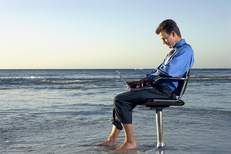 Businessman Sitting on a Beach by the Sea in a Swivel Chair and Working on His Laptop #1 Photograph by John Cumming