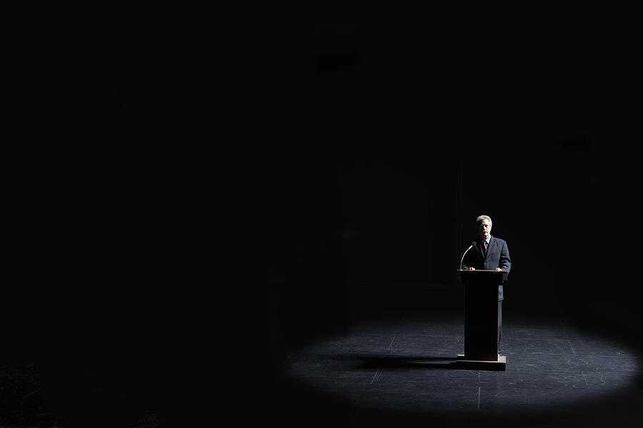 Businessman standing at podium illuminated by spotlight, elevated view #1 Photograph by Tom Fowlks