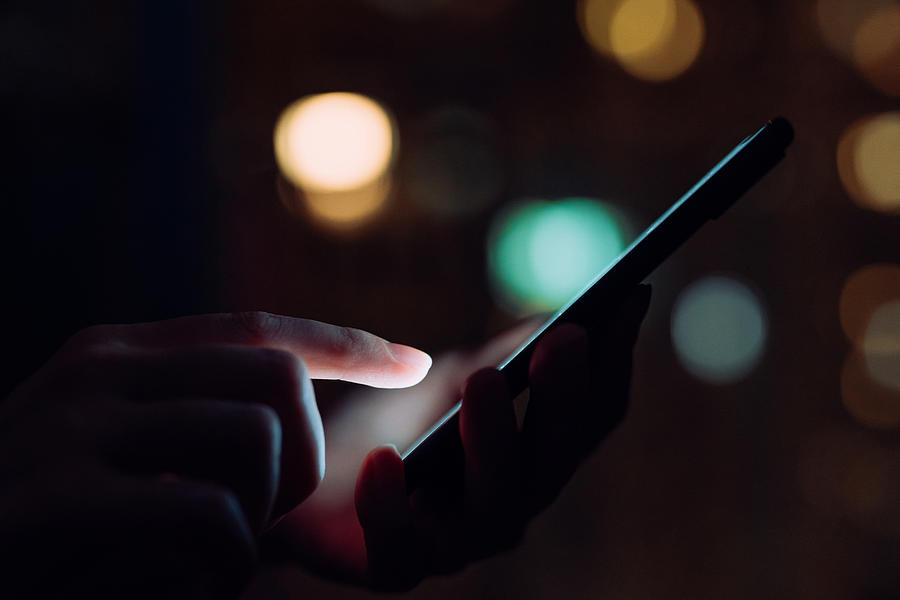 Close up of womans hand using smartphone in the dark, against illuminated city light bokeh #1 Photograph by D3sign