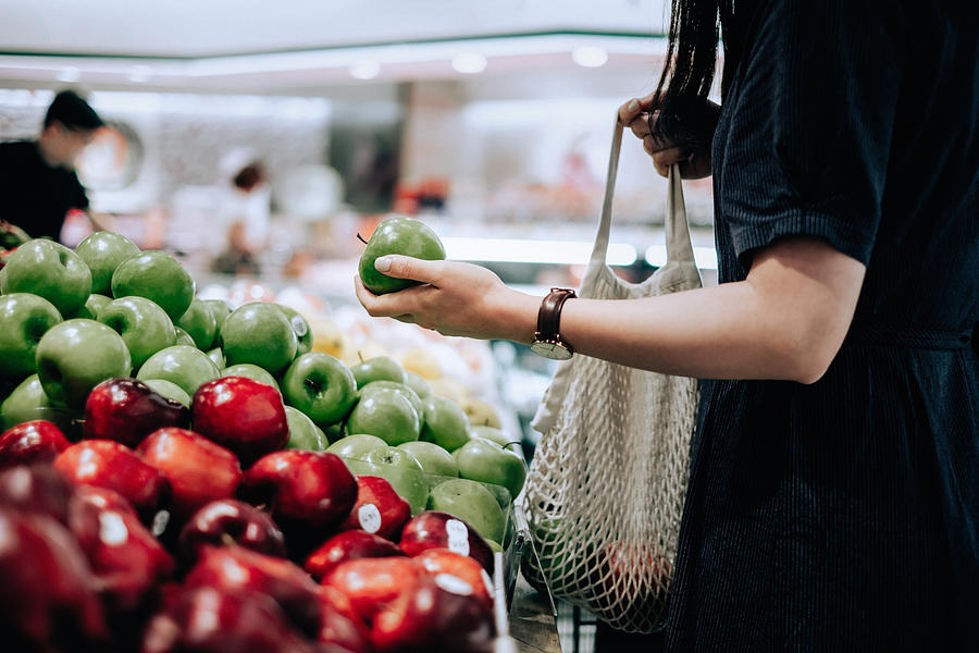 Cropped shot of young Asian woman shopping for fresh organic groceries in supermarket. She is shopping with a cotton mesh eco bag and carries a variety of fruits and vegetables. Zero waste concept Photograph by D3sign
