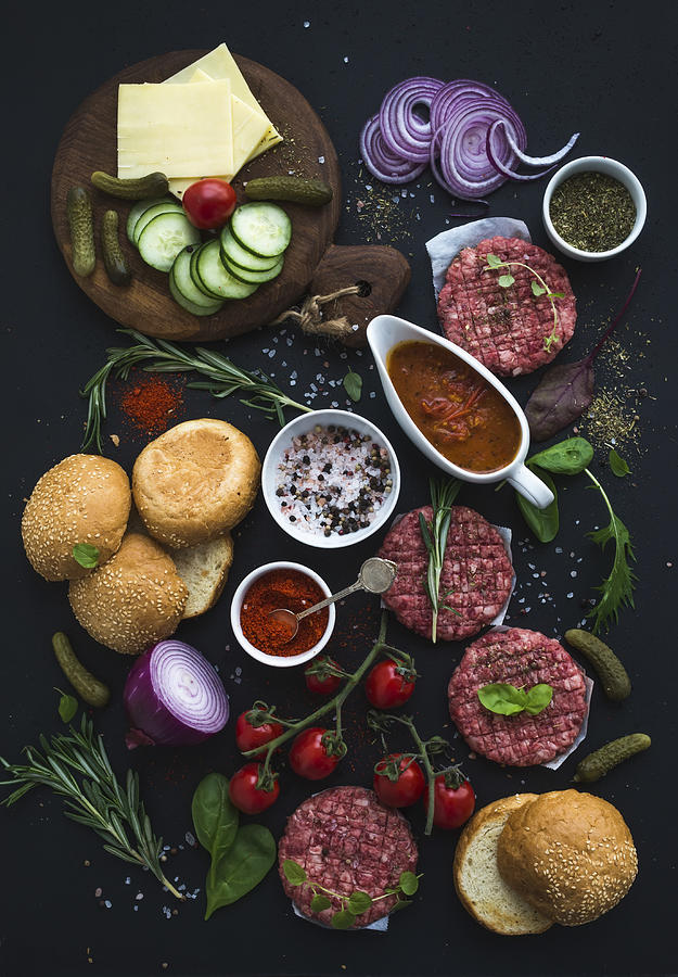 Ingredients for cooking burgers. Raw ground beef meat cutlets, buns, red onion, cherry tomatoes, greens, pickles, tomato sauce, cheese, herbs and spices over black background, top view. #1 Photograph by The Picture Pantry