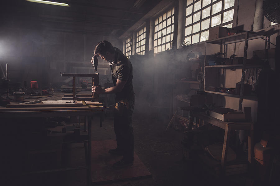 Male carpenter using drill to repair a chair in a workshop. #1 Photograph by Skynesher
