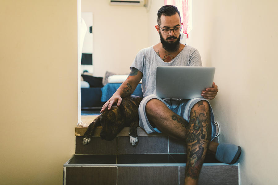 Man using laptop at home with his dog next to him #1 Photograph by Vgajic