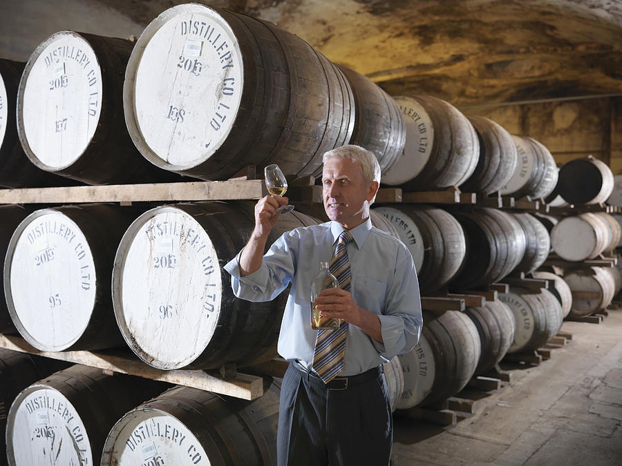 Manager with whisky sample next to ageing whisky barrels in distillery #1 Photograph by Monty Rakusen