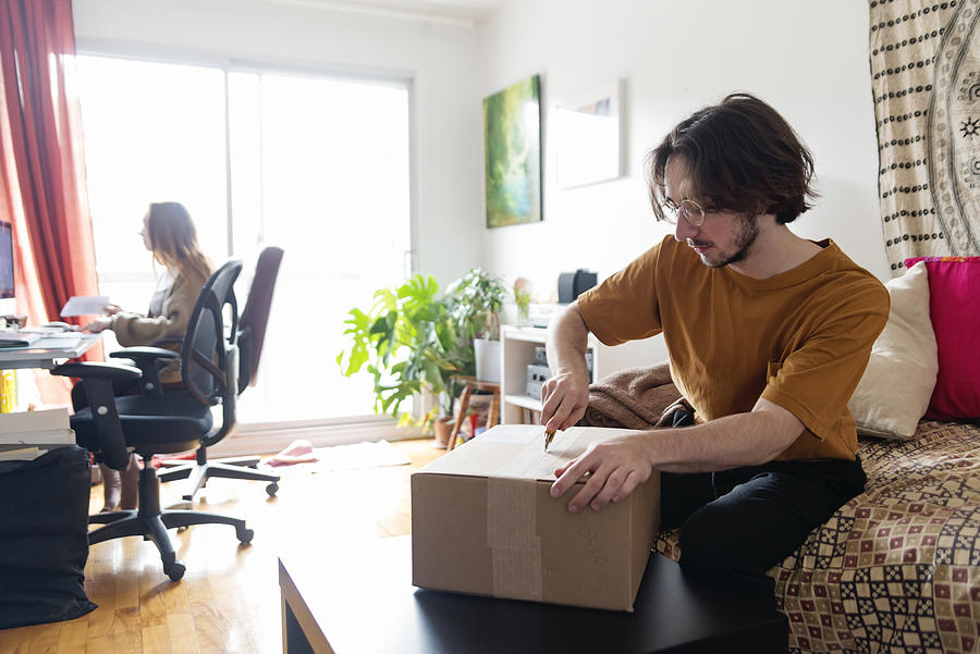 Millennial couple opening a delivered package in living room. #1 Photograph by Martinedoucet