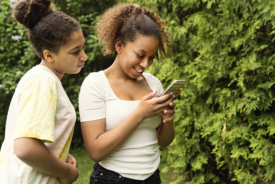 Mixed-race teenage sisters looking at mobile phone in backyard. #1 Photograph by Martinedoucet