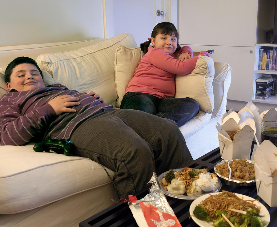 Overweight Brother and Sister Sitting on a Sofa Eating Takeaway Food and Watching the TV #1 Photograph by Digital Vision.