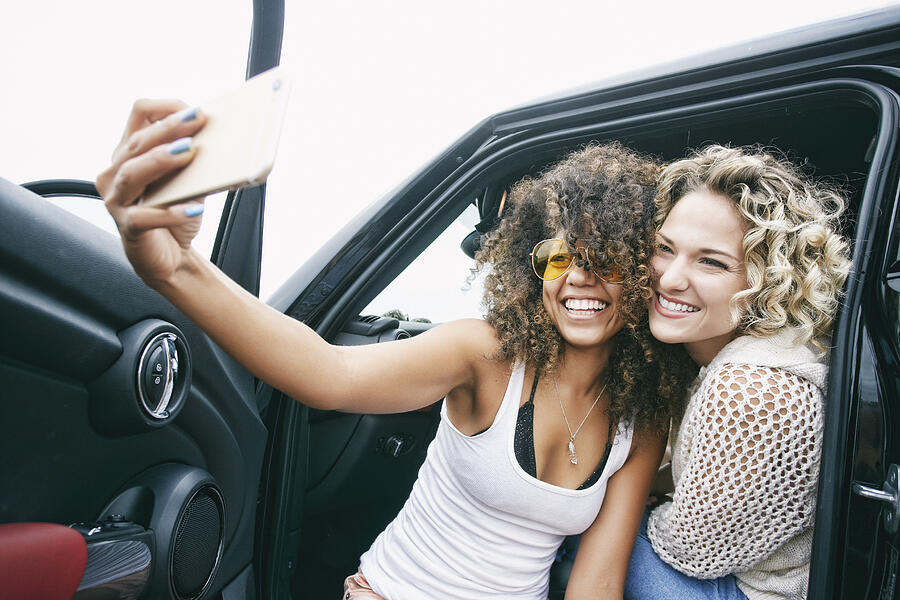 Portrait of two smiling young women with blond and brown curly hair sitting in car, taking selfie with mobile phone. #1 Photograph by Mint Images