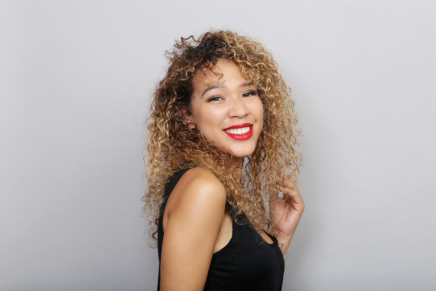Portrait of young LatinX woman with curly blonde hair, wearing red lipstick and a black tank top, in front of a grey background. #1 Photograph by Mireya Acierto