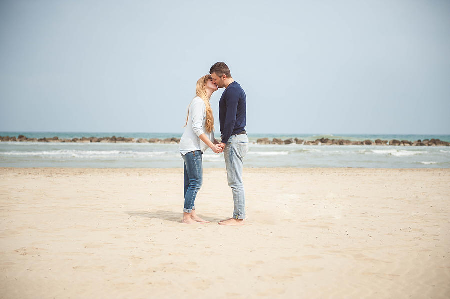 Pregnant couple kissing on the beach. Hand in hand. Casual clothes. #1 Photograph by © Samantha Carrirolo