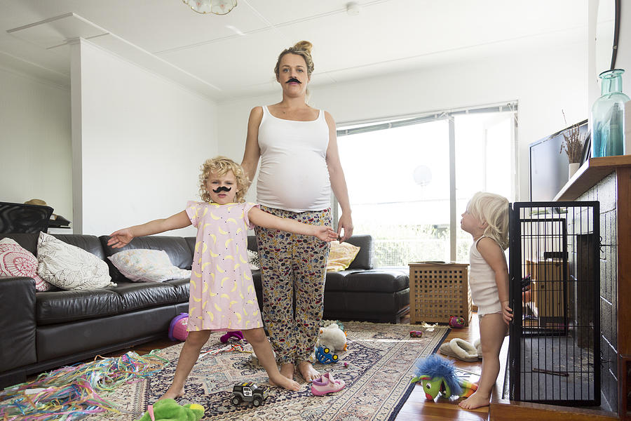 Pregnant mother and two children wearing moustaches in messy living room #1 Photograph by Jessie Casson