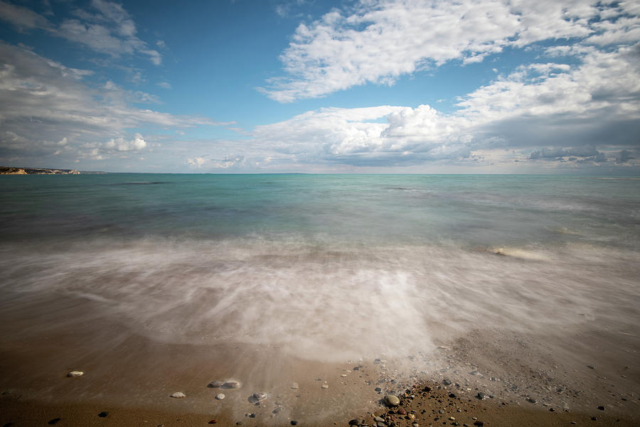 Seawaves splashing onto the sandy coast with pebbles and blue cloudy sky. #1 Photograph by Michalakis Ppalis