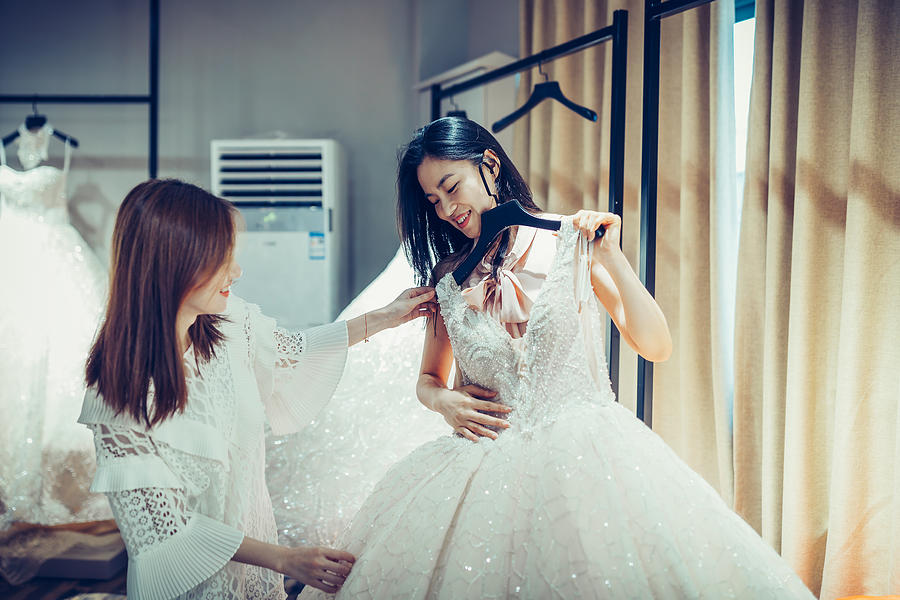 Two Asian Young women shopping for wedding dress gowns in boutique discount store, many white garments hanging on rack hangers row. #1 Photograph by Asia-Pacific Images Studio