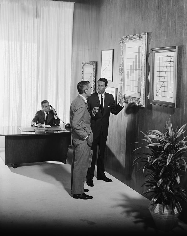 Two businessman discussing at bar chart while another man using telephone in background #1 Photograph by Tom Kelley Archive