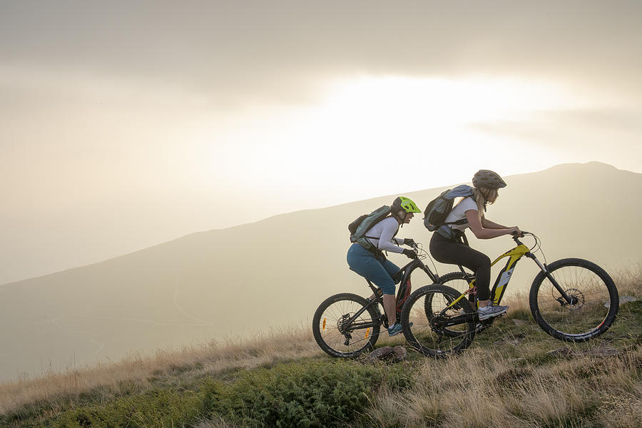 Two women ride up grassy hillside on electric mountain bikes #1 Photograph by AscentXmedia