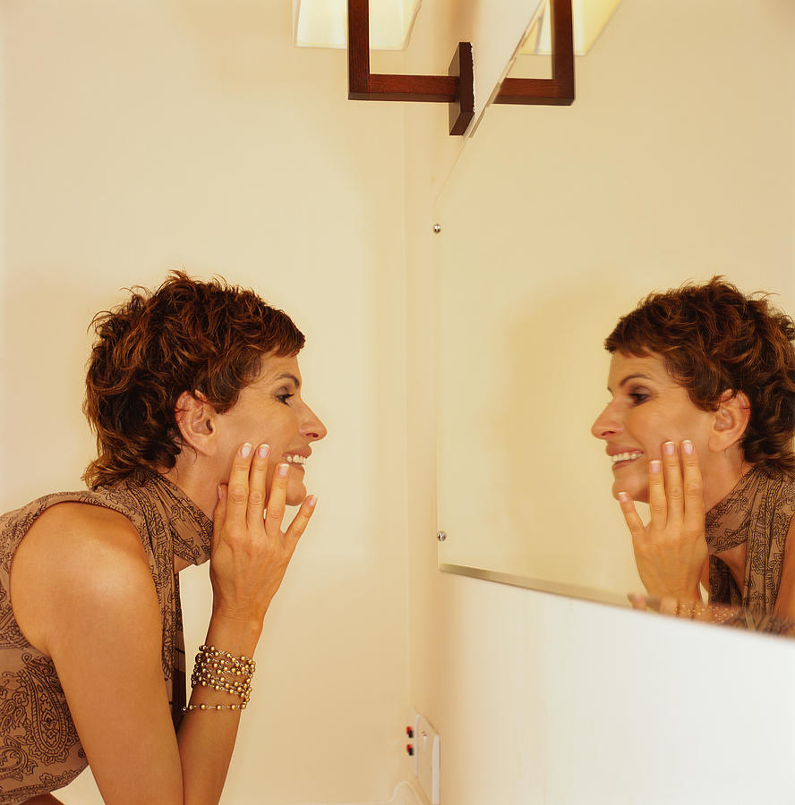 Woman Touching Her Face While Looking In The Mirror #1 Photograph by Stockbyte