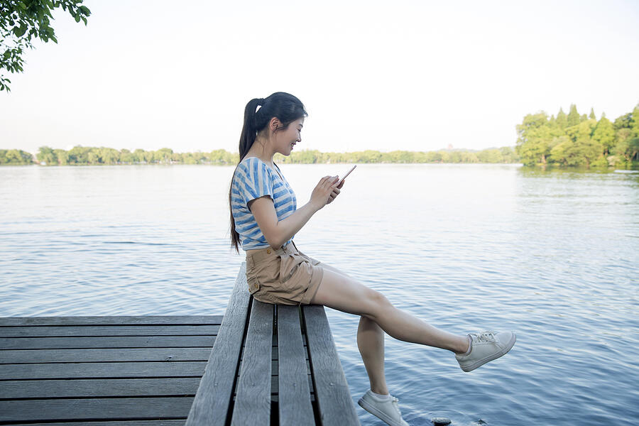 Young asian woman sitting on pier above lake,using smart phone #1 Photograph by Xia Yuan