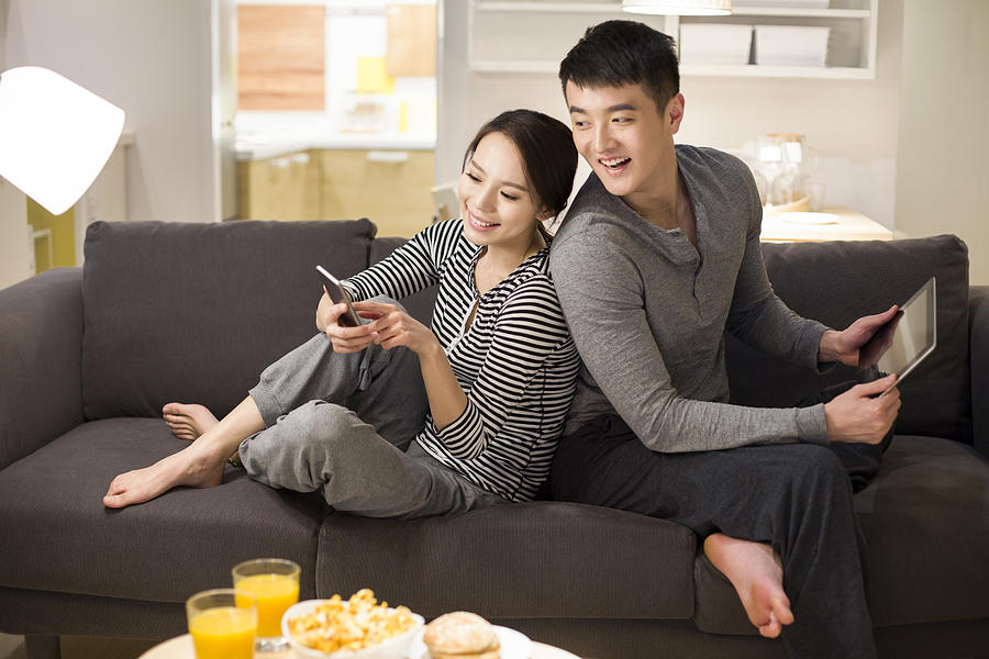 Young couple using smart phone on living room sofa #1 Photograph by BJI / Blue Jean Images