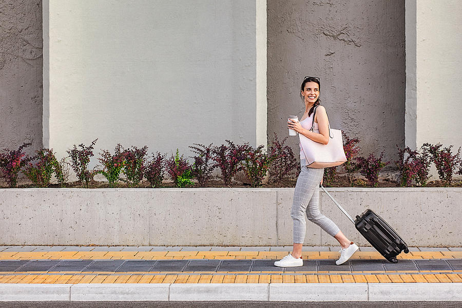 Young woman walking on a sidewalk beside the concrete wall and pulling a small wheeled luggage #1 Photograph by Gruizza