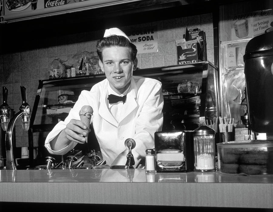 1950s Smiling Young Man Soda Jerk Leaning Across Counter Looking At Camera Serving Ice Cream Cone Photograph by Panoramic Images