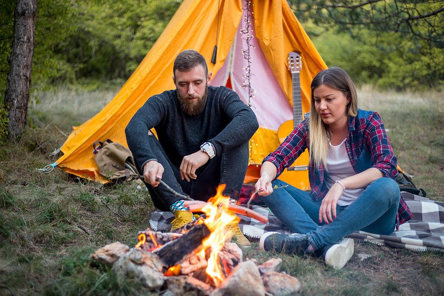 Beautiful couple, sitting, camping around the campfire. On the background with a tent The man flicks the fire with a stick. #2 Photograph by MajaMitrovic