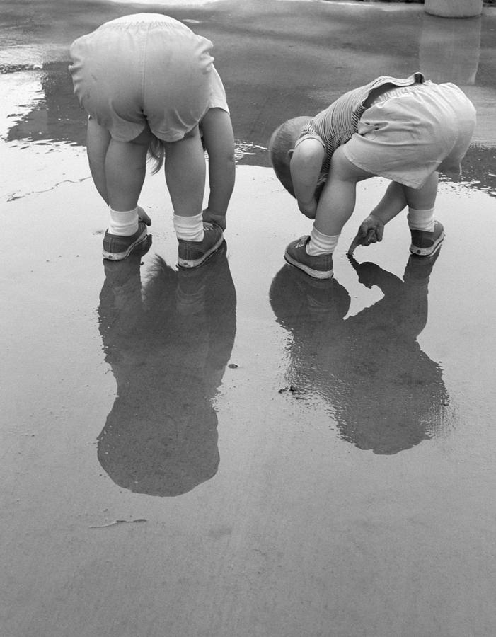 2 Kids Bending Over Rear View Backs To Camera Playing In Mud Puddle Rain Reflection In Water. Photograph by H. Armstrong Roberts