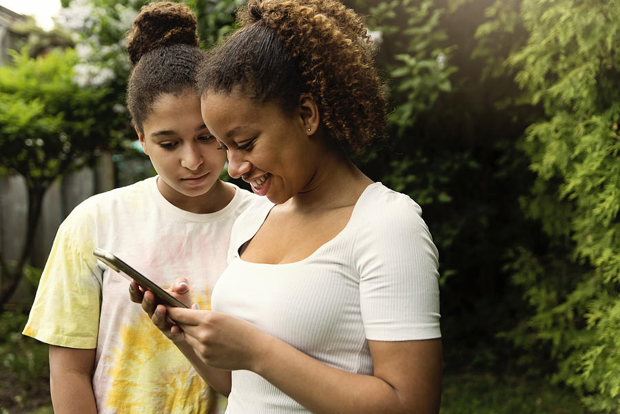 Mixed-race teenage sisters looking at mobile phone in backyard. #2 Photograph by Martinedoucet