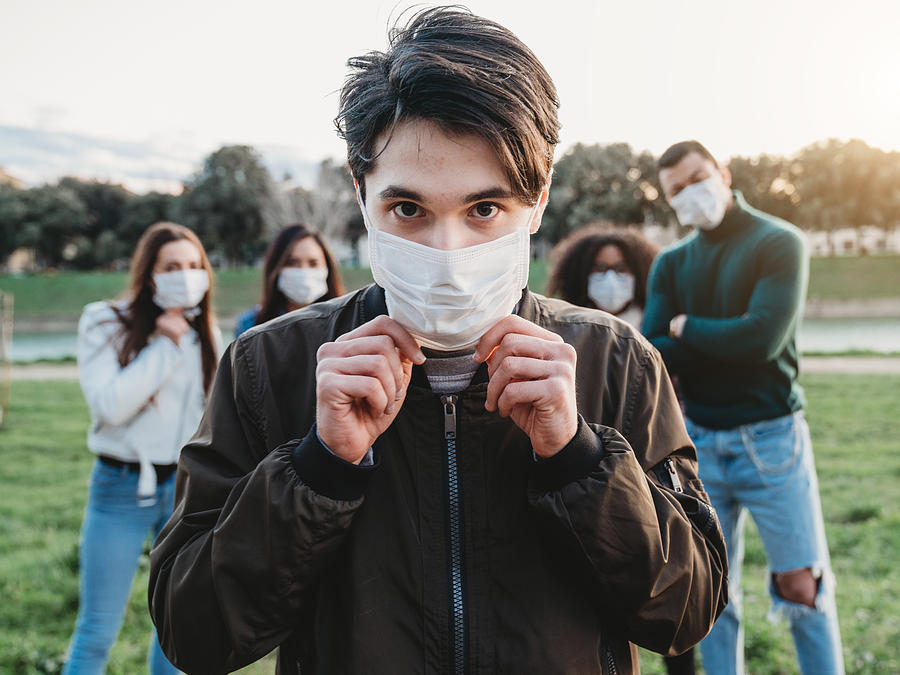 Young adult man wearing a pollution mask to protect himself from viruses. His friends are in the background. Photograph by FilippoBacci
