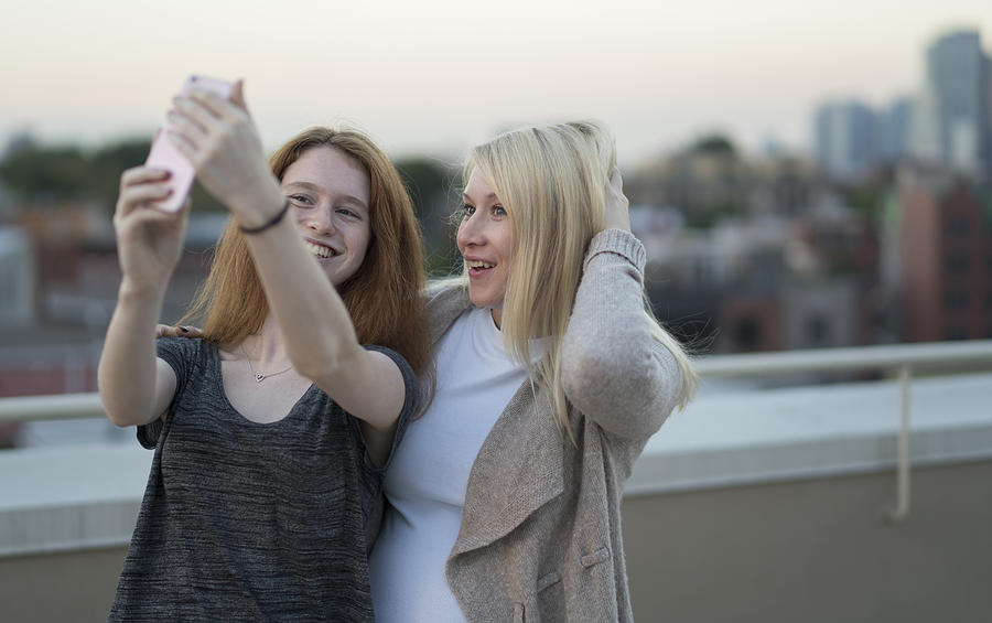 The young pretty pregnant woman with her little sister, the 16 years old teenager girl, hanging out together, taking selfies pictures with the smartphone and having fun at the rooftop #3 Photograph by Alex Potemkin