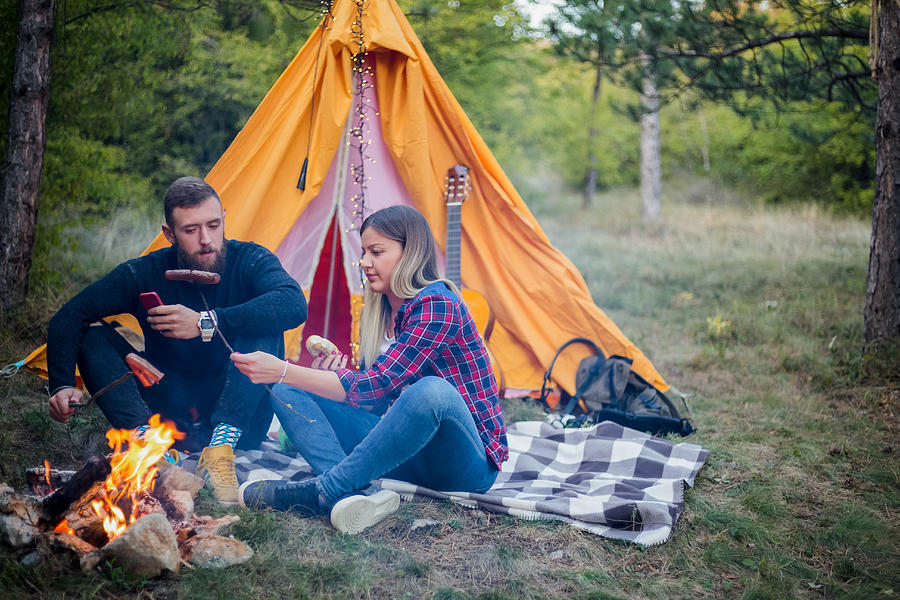 Beautiful couple, sitting, camping around the campfire. On the background with a tent The man flicks the fire with a stick. #9 Photograph by MajaMitrovic