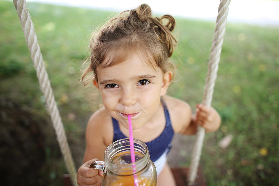 A 3 years old girl drinking orange juice seating on a swing Photograph by Catherine Delahaye