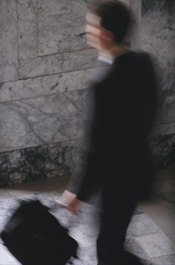 A blurred businessman in a black suit carries a brief case as he walks Photograph by Photodisc