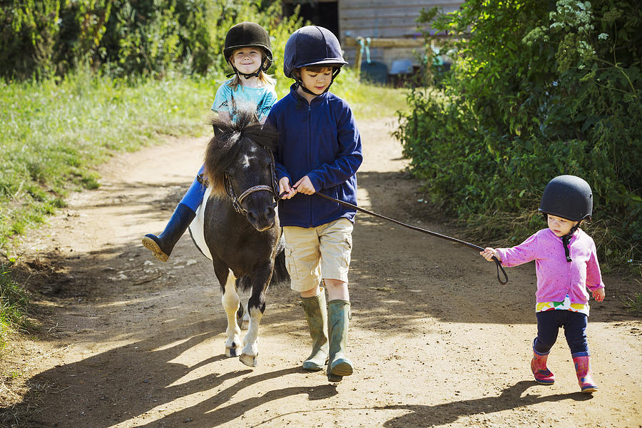 A boy, a toddler, and a girl riding a pony on a dirt path. Photograph by Mint Images