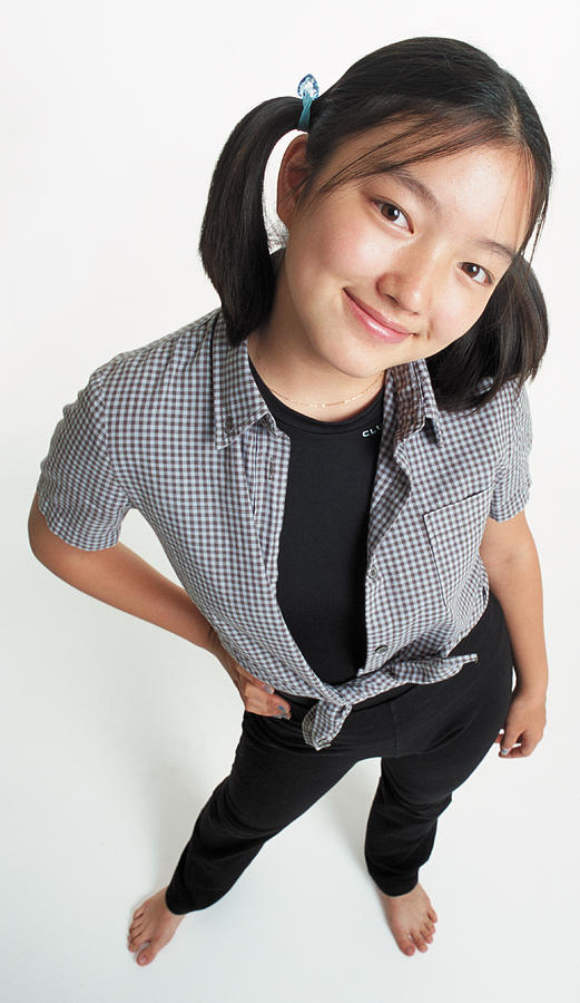 A Cute Young Asian Girl With Ponytails Wearing Black Stretch Pants With A Checkered Blouse Looks Up At The Camera Smiling With Her Head Cocked Photograph by Photodisc